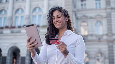 Junge Frau mit lockigen Haaren und Bluse steht vor Verwaltungsgebäude und vollzieht mit ihrer Bankkarte in der Hand zufrieden einen digitalisierten Verwaltungsprozess an ihrem Tablet