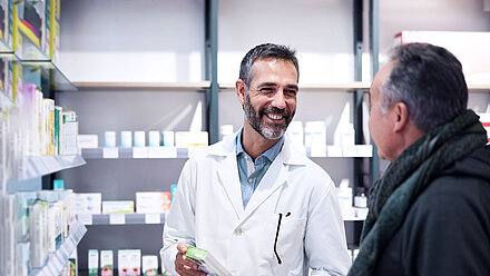 Zwei Männer in einer Apotheke; Apotheker mit kurzen, schwarzen Haare und Bart; trägt einen Arztkittel und hält ein Medikament in der Hand. Er lächelt einen Kunden an, der vor ihm steht, auch schwarze, kurze Haare, Jacke und Schal.